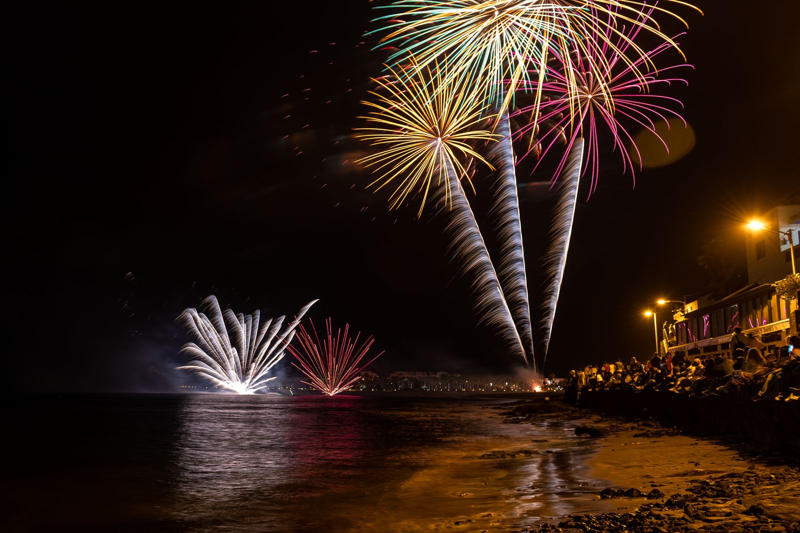 Feu d'artifice tiré depuis une plage la nuit, gerbes rouges et dorées au-dessus de la mer